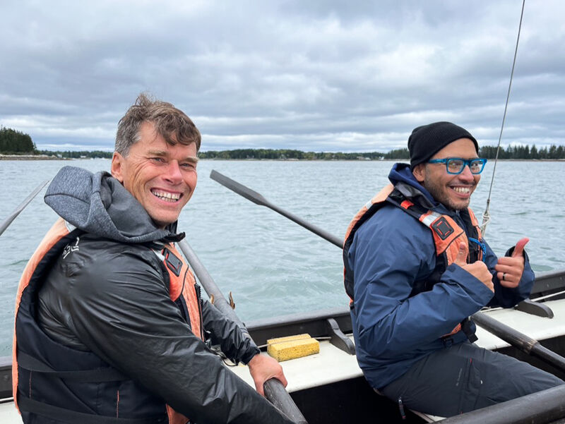 Two men are in a rowboat on a cloudy day. They are both wearing life jackets and smiling at the camera. One man is holding an oar, and the other is giving a thumbs up. The background shows a shoreline with trees.